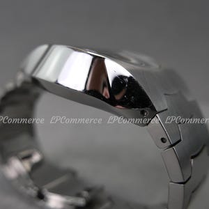 Op de afbeelding: Een close-up van een zilverkleurig polshorloge. Het horloge heeft een geometrisch ontwerp met een rechthoekige wijzerplaat en een bijpassende metalen band. Het horloge is schuin, met de zijkant en bovenkant zichtbaar.