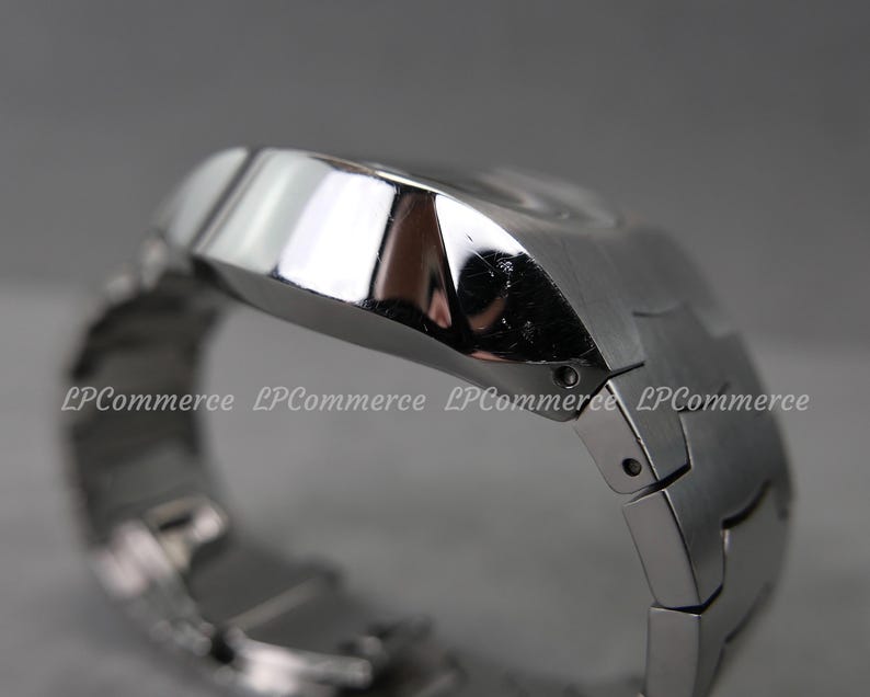 Op de afbeelding: Een close-up van een zilverkleurig polshorloge. Het horloge heeft een geometrisch ontwerp met een rechthoekige wijzerplaat en een bijpassende metalen band. Het horloge is schuin, met de zijkant en bovenkant zichtbaar.