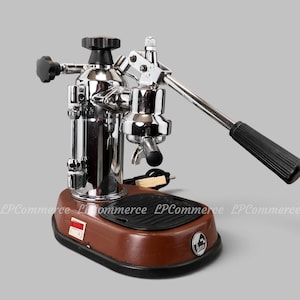 May include: A chrome and brown lever espresso machine featuring a vertical boiler, a long black-handled manual lever, a black drip tray, and a small rectangular power switch on the base. A circular logo with the text La Pavoni is visible.