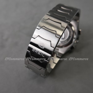 Op de afbeelding: Een close-up van een zilverkleurig Breil horloge. De horlogeband is gemaakt van in elkaar grijpende metalen schakels. De merknaam "Breil" is zichtbaar op de band. De wijzerplaat van het horloge is gedeeltelijk zichtbaar op de achtergrond.
