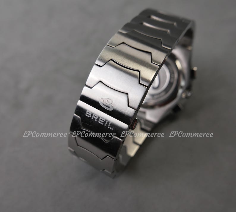 Op de afbeelding: Een close-up van een zilverkleurig Breil horloge. De horlogeband is gemaakt van in elkaar grijpende metalen schakels. De merknaam "Breil" is zichtbaar op de band. De wijzerplaat van het horloge is gedeeltelijk zichtbaar op de achtergrond.