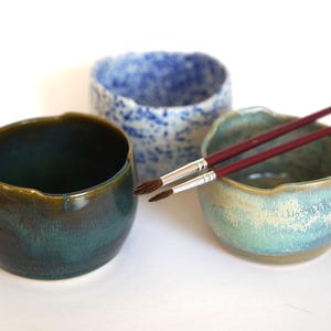 May include: Three ceramic bowls, one blue and white, one green, and one light blue and beige, with two paintbrushes resting on the rim of the light blue bowl.