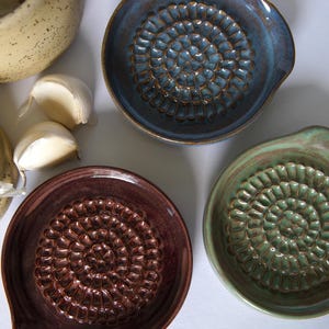 May include: Three ceramic garlic presses in different colors: blue, green, and brown. Each press has a textured surface with a circular pattern.