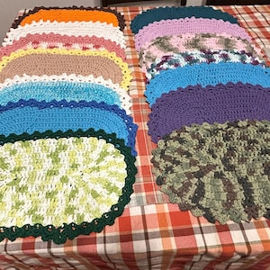 May include: A collection of colorful, oval-shaped crochet placemats. The placemats feature a scalloped edge and are in various colors, including orange, white, pink, yellow, brown, blue, green, purple, and camouflage.