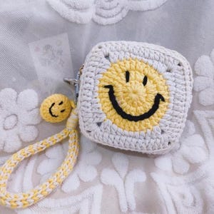 Op de afbeelding: Een witte en gele gehaakte portemonnee met een lachend gezicht ontwerp. De portemonnee heeft een witte gehaakte band met een gele gehaakte smiley-hanger.