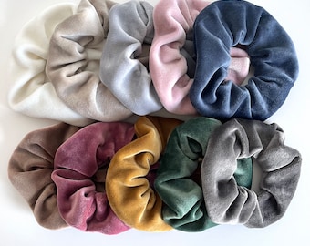 Può includere: Una collezione di dieci elastici per capelli in velluto di vari colori, tra cui crema, beige, grigio, rosa, blu navy, marrone, bordeaux, oro, verde e antracite. Questi accessori per capelli sono disposti su una superficie bianca.