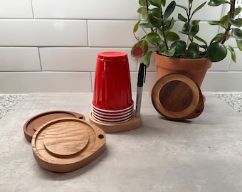 May include: Wooden coasters with a natural finish, paired with a red plastic cup and a black marker. The coasters have a circular design with a raised center and a small handle. A potted plant is in the background.