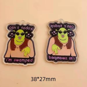 Puede incluir: Dos colgantes acrílicos con un personaje de dibujos animados verde con gafas de sol. Los colgantes tienen el texto "can't today, I'm swamped" y "38*27mm". El personaje lleva un chaleco marrón y una camisa rosa claro.