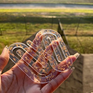 以下が含まれることがあります： 金箔が埋め込まれた透明なアーチ型のコースター。 コースターは手で持たれており、背景はぼやけた緑と茶色です。 ユニークでモダンなデザインです。