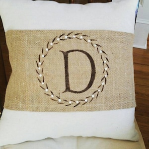 May include: A decorative pillow with a white background and a burlap band. The burlap band features a brown letter "D" inside a wreath design. The wreath is made of brown leaves and white dots.