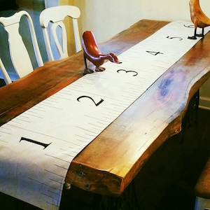 May include: A white table runner with black numbers 1 through 5 printed on it. The runner is laid out on a wooden table with a wooden duck figurine on top of it.