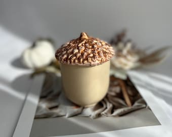 May include: A ceramic acorn-shaped container with a beige base and a textured, brown cap. The container is on a magazine with the text "THE MAGAZINE" and the phrase "Live well, love lots, and laugh often."