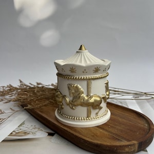 May include: A carousel-themed trinket box in off-white with gold detailing. The design features carousel horses and star motifs. The conical lid and the box are presented on a wooden tray.