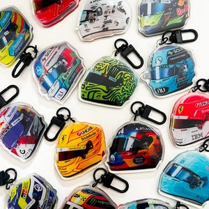 May include: A collection of thirteen acrylic keychains shaped like Formula 1 racing helmets, each featuring distinct team colors, sponsor logos, and visor designs. Each keychain includes a black plastic swivel clasp for attachment.