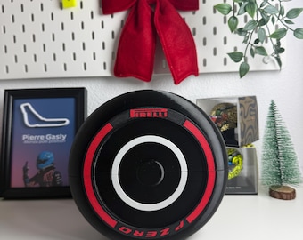 Op de afbeelding: Een zwart Pirelli P Zero Formule 1-bandmodel met een opvallende rode streep rond de omtrek, een wit cirkelvormig detail in het midden en het Pirelli-logo in rode letters op de bovenkant van de zijkant van de band.