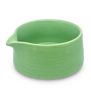 May include: A light green ceramic creamer or small pouring bowl with a smooth, glossy finish and a subtle horizontal ribbed texture along the exterior. The vessel features a distinct, integrated spout on one side for controlled pouring.