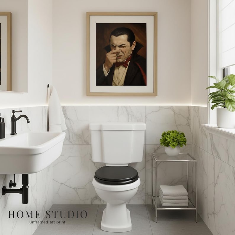 May include: A framed art print featuring a portrait of a vampire in a black suit, red bow tie, and high-collared cape, holding their nose with one hand. The print is displayed in a light wood frame on a white wall above a white toilet.