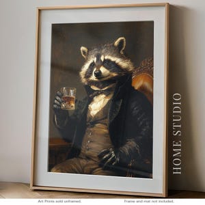 May include: Framed art print featuring a raccoon dressed in formal attire, holding a glass of amber liquid. The raccoon is seated in a leather chair, with a dark background. The print is in a gold frame with the text "HOME STUDIO" on the side.
