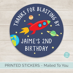 Op de afbeelding: Ronde blauwe en witte sticker met een rood raketje, planeten en sterren. De tekst luidt "Bedankt voor het opstijgen met Jaime voor zijn 2e verjaardag".