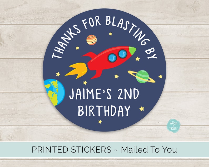 Op de afbeelding: Ronde blauwe en witte sticker met een rood raketje, planeten en sterren. De tekst luidt "Bedankt voor het opstijgen met Jaime voor zijn 2e verjaardag".