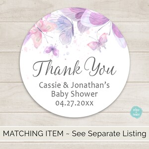 Op de afbeelding: Ronde bedank-sticker met een aquarelontwerp van paarse en roze vlinders op een witte achtergrond. De tekst "Bedankt" staat in een grijs handschrift. Onder de tekst staat "Baby Shower van Cassie & Jonathan 04.27.20XX".