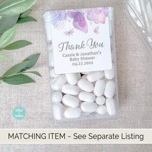 Op de afbeelding: Een doorzichtige plastic container gevuld met witte pepermuntjes. Een klein bedankkaartje met paarse vlinders en de tekst "Thank You Cassie & Jonathan's Baby Shower 04.27.20XX" is bevestigd aan de container.