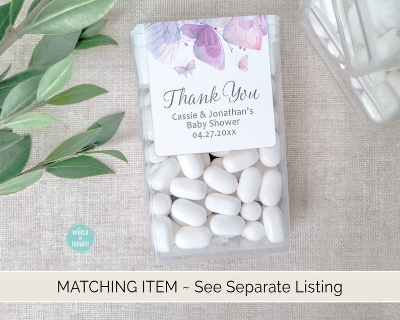 Op de afbeelding: Een doorzichtige plastic container gevuld met witte pepermuntjes. Een klein bedankkaartje met paarse vlinders en de tekst "Thank You Cassie & Jonathan's Baby Shower 04.27.20XX" is bevestigd aan de container.