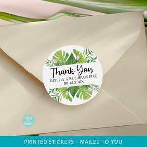 May include: A circular white sticker featuring a watercolor tropical leaf design in shades of green with small pink accents. The text reads Thank You, GISELLE'S BACHELORETTE, and 08.14.20XX. The sticker is centered on a light beige envelope.