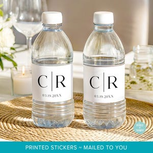 May include: Two clear plastic water bottles with white caps, each featuring a white rectangular label. The labels display the black serif letters C and R separated by a vertical line, with the date 03.19.20XX printed in black below the letters.