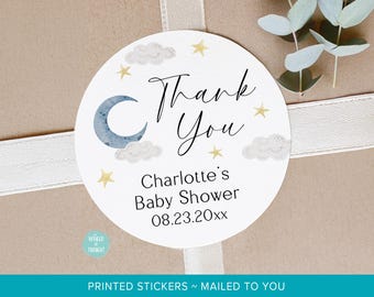 Könnte beinhalten: Ein runder weißer Aufkleber mit einer blauen Mondsichel, grauen Wolken und gelben Sternen. Der Text lautet Thank You, Charlotte's Baby Shower und 08.23.20xx. Der Aufkleber hat eine glatte Oberfläche und liegt auf einem hellbraunen Untergrund mit Band.