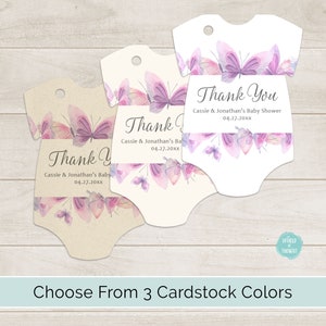 Op de afbeelding: Drie baby-rompertjes in de vorm van bedankkaartjes met een aquarel-vlinderontwerp in tinten paars en roze. De tekst op de kaartjes luidt "Bedankt" met de naam "Baby Shower van Cassie & Jonathan" en de datum "04.27.20xx".