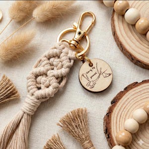 May include: A beige macrame keychain with a gold-colored clasp and a wooden tag. The tag has etched text and a drawing. The keychain has a woven design and a tassel. Decorative elements include wooden beads and dried plants.