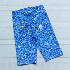 May include: Blue glitter print biker shorts. The shorts have a high waist and are made of a stretchy material. The text "Little Darling" is printed on the shorts.