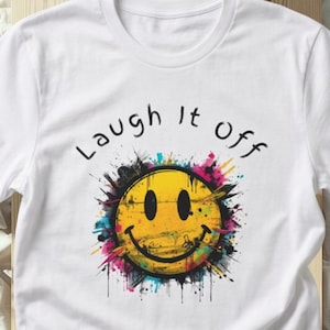 May include: A white short-sleeved t-shirt featuring a central graphic of a yellow smiley face surrounded by colorful paint splatters in shades of pink, blue, and black. The text "Laugh it off" is printed in black above the smiley face design.
