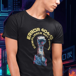以下が含まれることがあります： ロボットアームが拳を作っている様子のスタイリッシュなイラストが描かれた黒いTシャツ。グラフィックには、黄色の円形デザインで「ERROR 404: COMPLIANCE NOT FOUND」というテキストが含まれています。Tシャツは人が着ています。