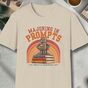 May include: Beige t-shirt with a retro design featuring a robot standing on books, with the text "Majoring in Prompts" above and "Minor in Deadlines" below. The design includes a rainbow arch.