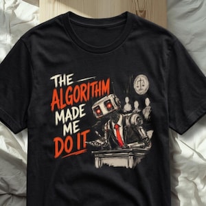 May include: A black t-shirt featuring a graphic of a robot in a suit and red tie standing at a podium. The text reads THE ALGORITHM MADE ME DO IT in red and white lettering. A circular icon with a scale of justice is visible in the background.