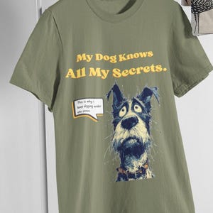 May include: Olive green t-shirt with the text "My Dog Knows All My Secrets." A cartoon dog with a surprised expression is featured below the text. The dog has blue and black fur and is wearing a collar. A speech bubble says "This is why..."