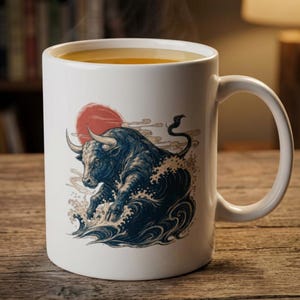 May include: A white ceramic mug filled with a golden liquid, featuring a graphic illustration of a dark blue bull emerging from stylized ocean waves beneath a large red sun circle and light-colored cloud accents on the side.