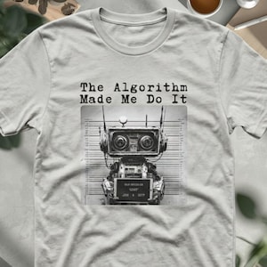 May include: A light gray t-shirt featuring a black and white graphic of a robot standing in front of a height chart. The robot holds a sign, and the text above the graphic reads, "The Algorithm Made Me Do It" in a typewriter-style font.