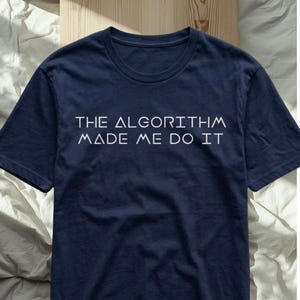 May include: A navy blue short-sleeved t-shirt featuring the white text THE ALGORITHM MADE ME DO IT printed in a geometric sans-serif font across the chest. The shirt has a crew neckline and a standard fit, showcasing a clean and simple design.