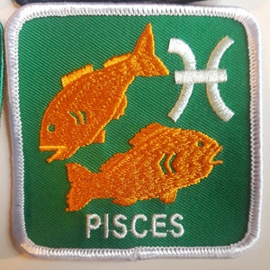 Könnte beinhalten: Ein grüner Stoff-Patch mit weißem Rand und dem Wort "PISCES" in weiß gestickt. Zwei orangefarbene Fische sind auf dem Patch gestickt, zusammen mit einem weißen astrologischen Symbol für Fische.