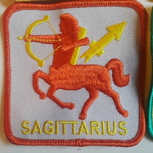 May include: A white embroidered patch with an orange and yellow image of a centaur holding a bow and arrow. The text "SAGITTARIUS" is embroidered in yellow below the image.