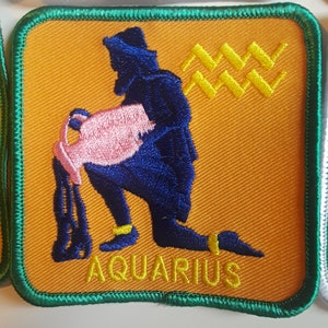 May include: A yellow and green embroidered patch with a blue figure pouring water from a pink pitcher. The figure is kneeling and the patch says "AQUARIUS" in yellow letters.