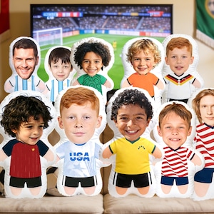 Peut inclure: Plusieurs coussins en peluche en forme de personnes, chacun avec un visage différent et portant des tenues sur le thème du football. Les coussins représentent diverses nationalités, dont les États-Unis, l'Allemagne et l'Argentine. Un match de football est visible à la télévision.