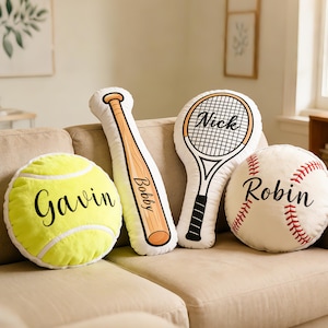 Puede incluir: Cuatro cojines decorativos con temática deportiva sobre un sofá beige. Los cojines incluyen una pelota de tenis amarilla con "Gavin", un bate de béisbol con "Bobby", una raqueta de tenis con "Nick" y una pelota de béisbol con "Robin".