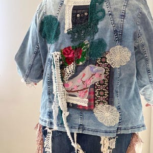 Può includere: Giacca di jeans blu chiaro con toppe decorative. Le toppe includono elementi floreali, in pizzo e all'uncinetto nei toni del rosa, verde e bianco. La giacca ha un aspetto unico e fatto a mano.