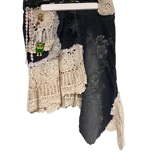 Può includere: Gonna di jeans scura con finiture e dettagli in pizzo crochet color crema. La gonna presenta ricami floreali, una piccola applicazione a forma di gufo e abbellimenti di perline. La gonna è appesa a una gruccia bianca.