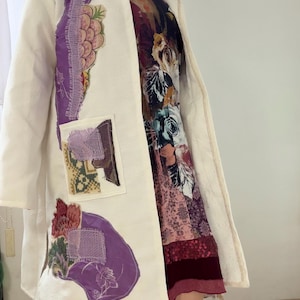 Könnte beinhalten: Ein cremefarbener Mantel mit Kapuze und lila und floralen Applikationen. Der Mantel hat einen strukturierten Stoff und ein Patchwork-Design. Der Mantel ist offen und zeigt ein floral gemustertes Kleid darunter. Der Mantel ist knielang.