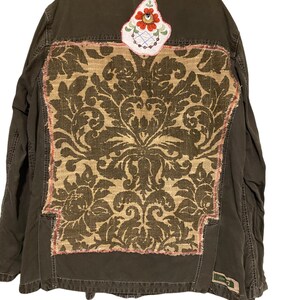 Könnte beinhalten: Dunkel olivgrüne Jacke mit einem großen Jute-Panel mit einem Damastmuster in Braun- und Beigetönen. Auf dem oberen Rücken ist ein kleiner, gestickter Blumenaufnäher aufgenäht. Die Jacke hat eine Knopfleiste und mehrere Taschen.